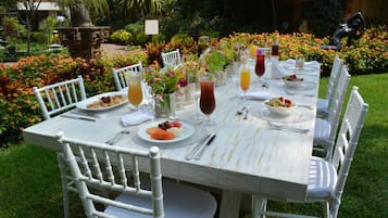 An outdoor dining table set with white chairs, surrounded by a garden with flowers and a statue.