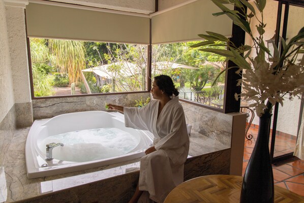 A person sitting in a bathtub with a view of a garden through a window. - Hostería Las Quintas Hotel (Cuernavaca)