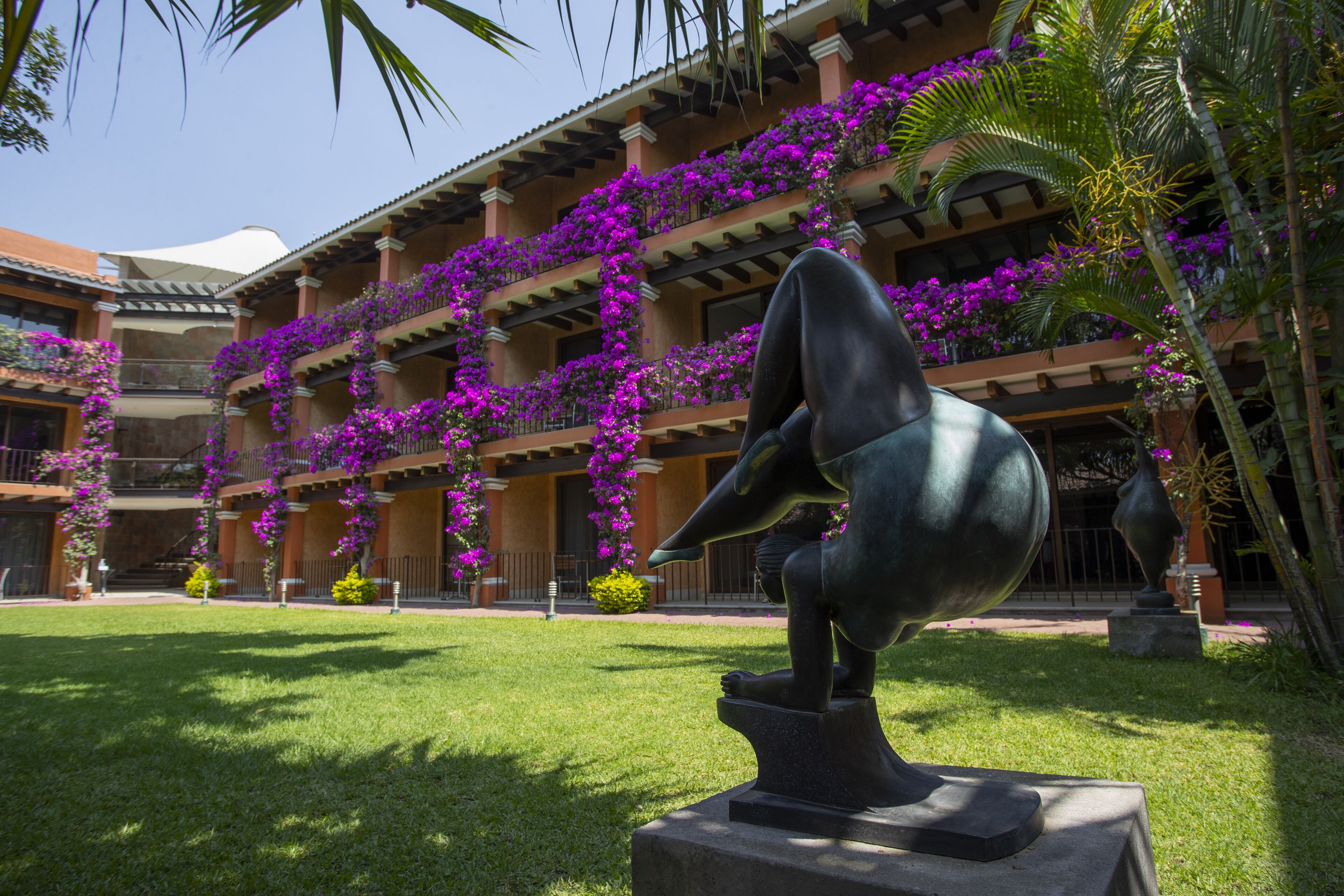 A courtyard with a sculpture, a building with balconies covered in purple flowers, and a palm tree.