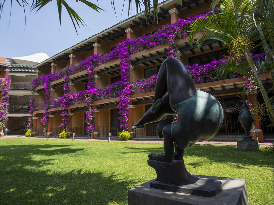 Un patio con una escultura, un edificio con balcones cubiertos de flores moradas y una palmera.