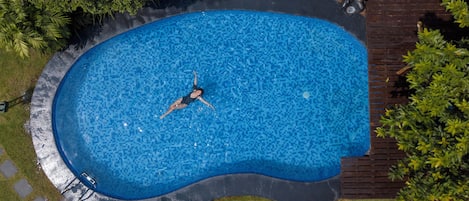 An aerial view of a swimming pool with a person swimming, surrounded by greenery and a wooden deck.