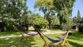 Una hamaca montada sobre un soporte de madera en un jardín bien cuidado, con varios árboles y un cielo despejado.