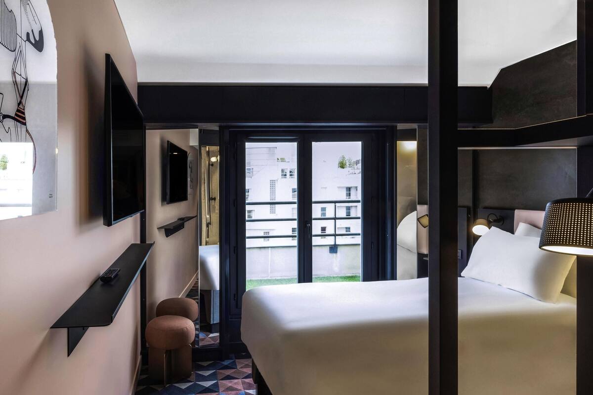A modern hotel room with a large bed, a TV, a small seating area, and a view of the outside.