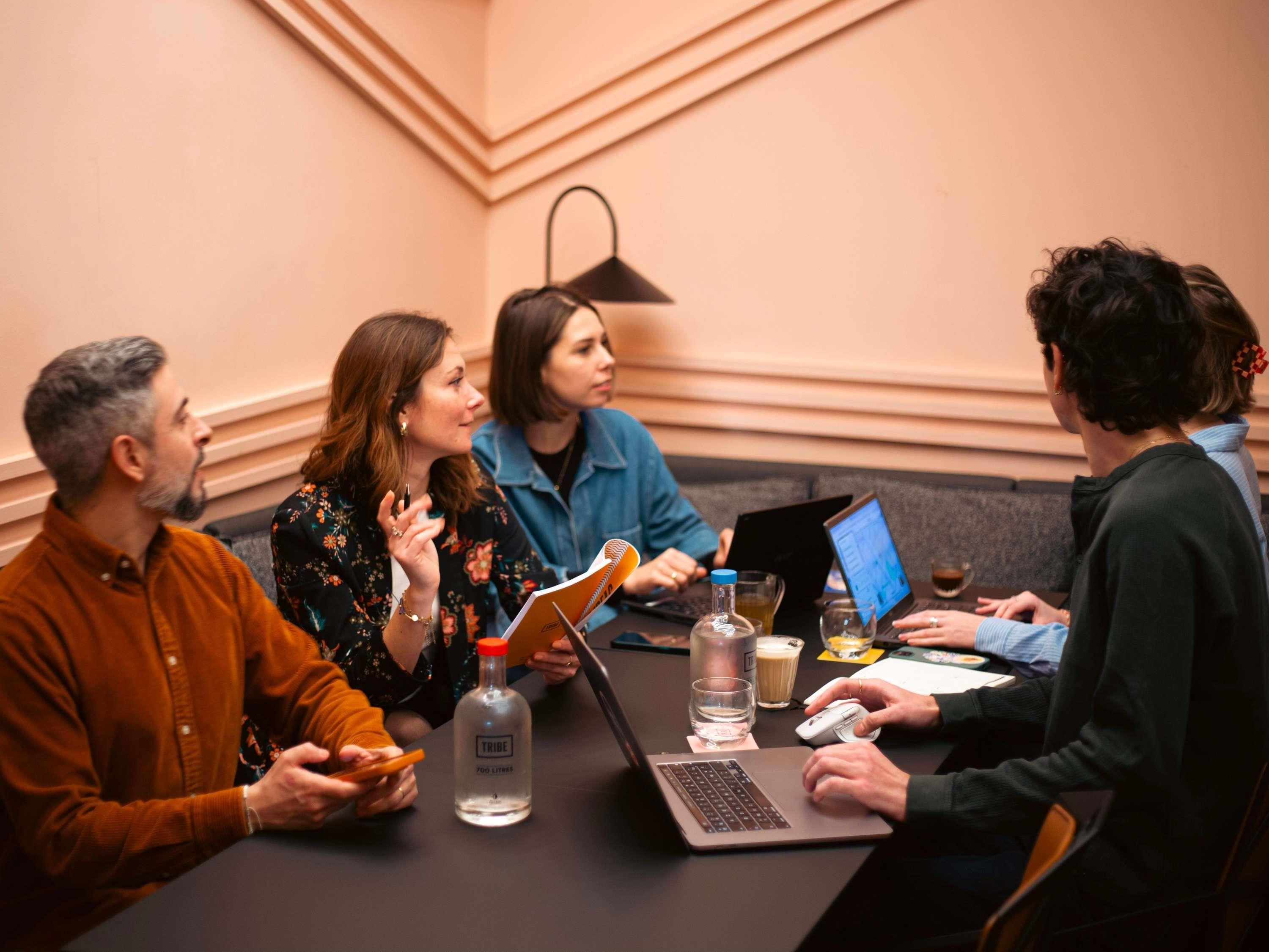 A group of people seated around a table in a conference room with laptops, a bottle of water, and a notebook.