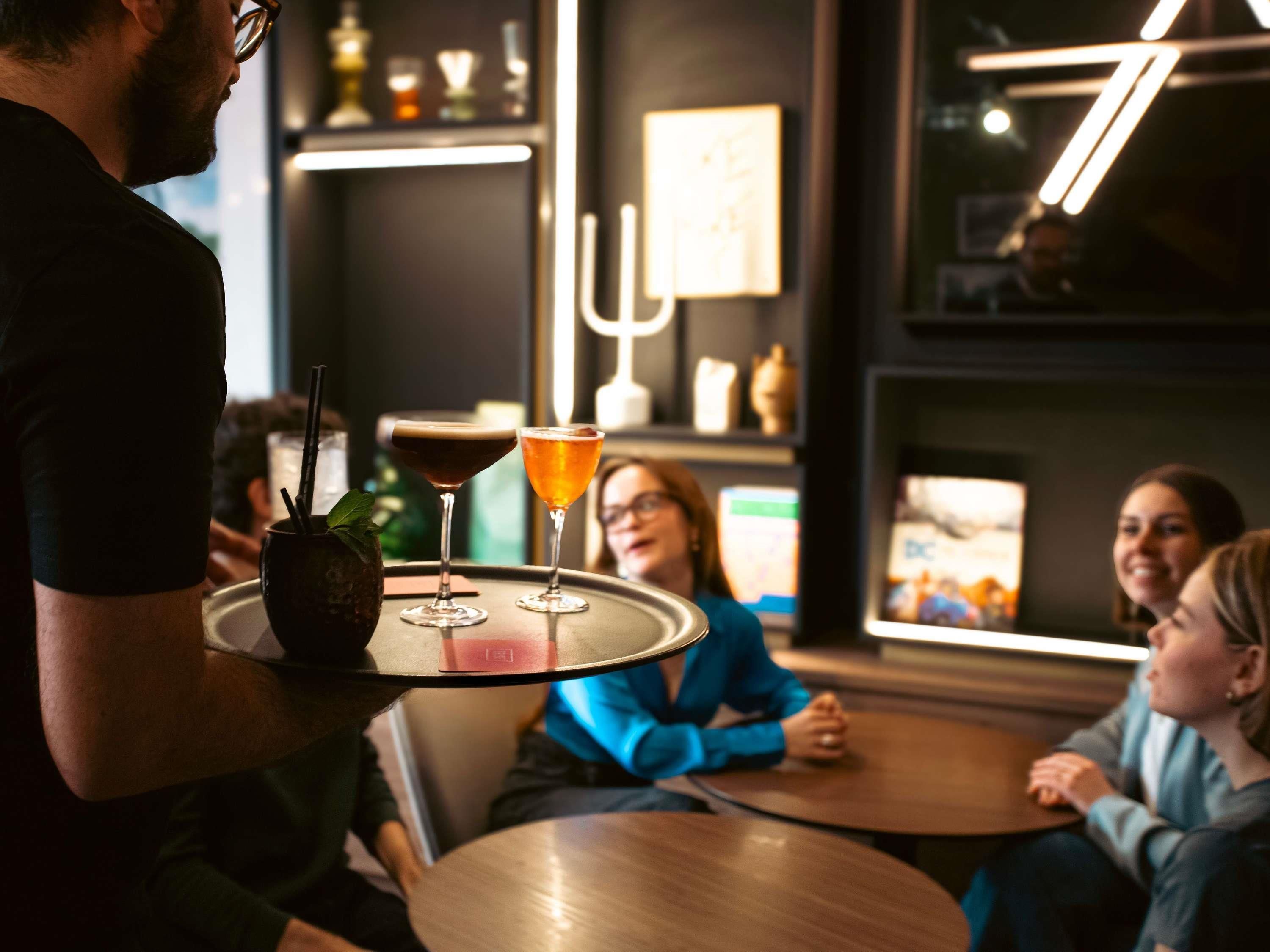 A bar setting with a waiter carrying a tray with two drinks, a bar counter, and a wall with decorative items.