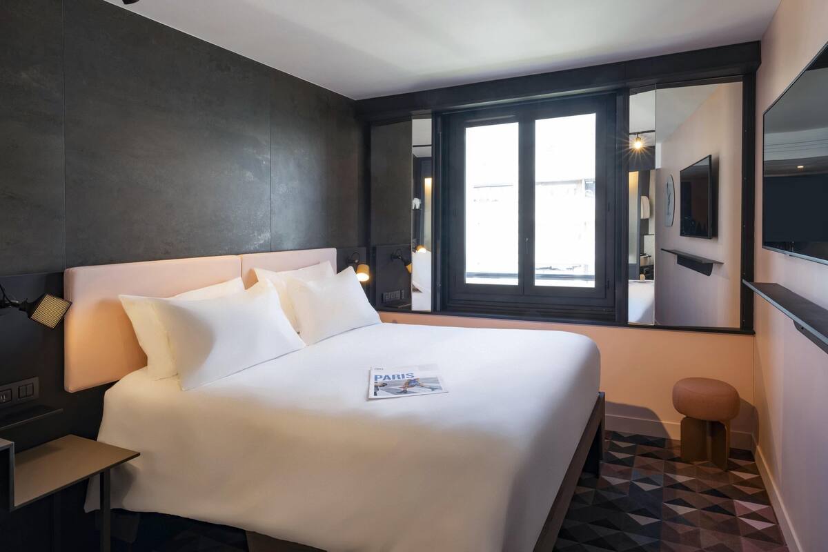 A modern hotel room with a large bed, a bedside table, a nightstand, a small stool, a window with a view, and a wall-mounted TV.
