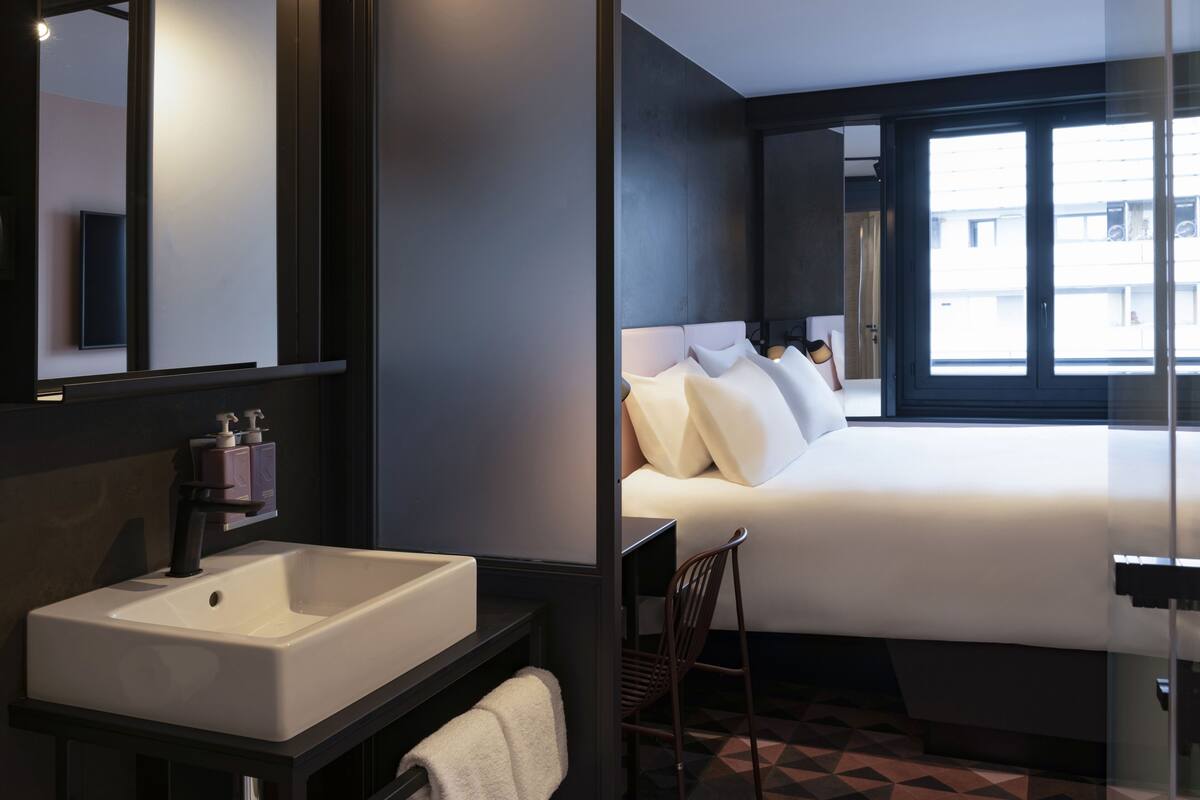 A modern hotel room with a large bed, a bedside table, a mirror, and a bathroom with a sink and towel rack.