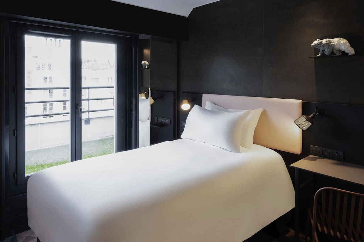 A hotel room with a large bed, a desk with a chair, a small shelf with a polar bear figurine, and a view through a sliding door.