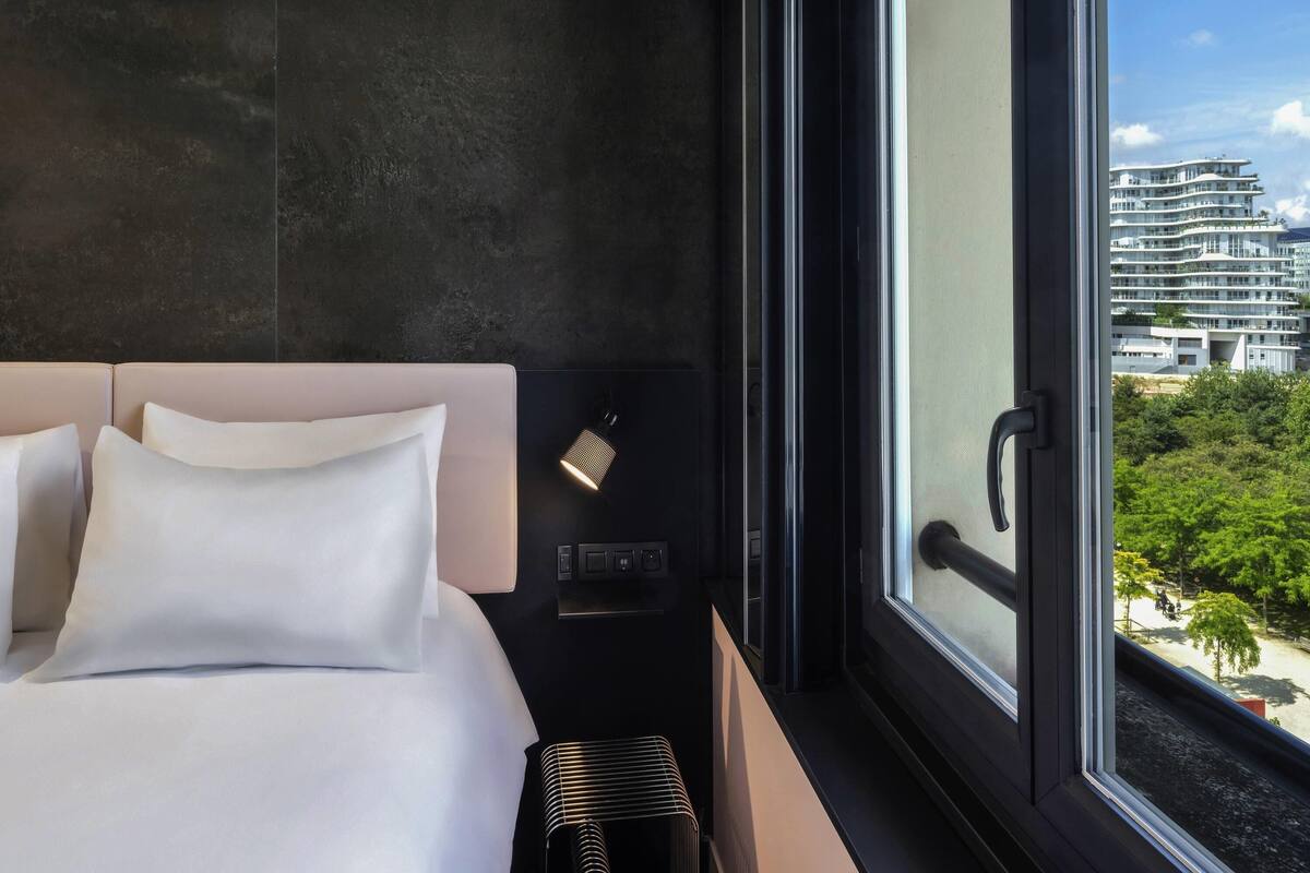 A modern hotel room with a large bed, a nightstand, a lamp, and a view of a cityscape through a large window.
