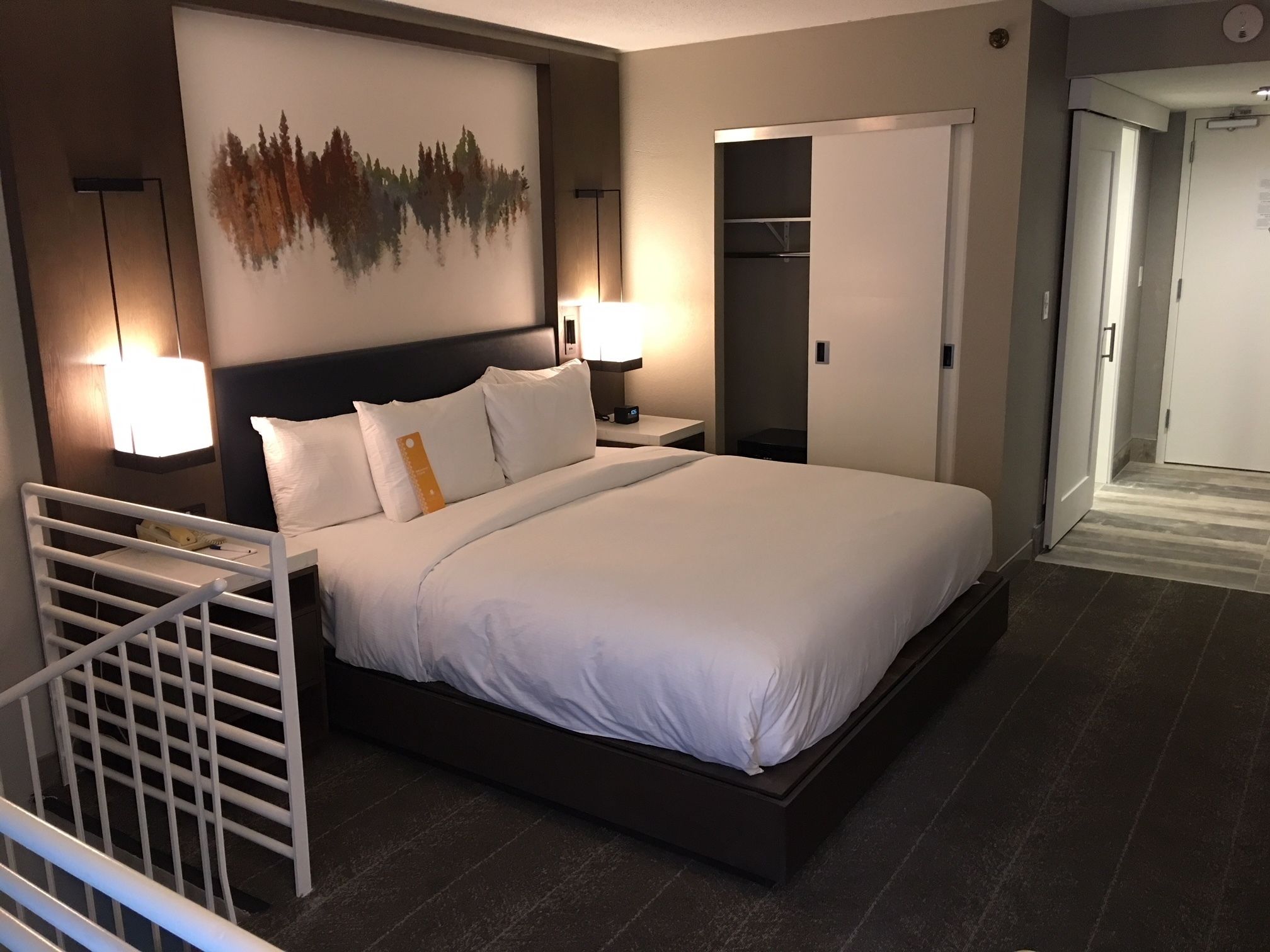 Una habitación de hotel moderna con una cama grande, dos lámparas de noche, una mesita de noche, un armario y una obra de arte colgada en la pared.