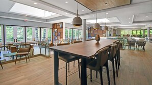 Un espacio interior amplio con una gran mesa y sillas de madera, un techo alto con tragaluces y ventanas grandes que ofrecen vistas a la vegetación.