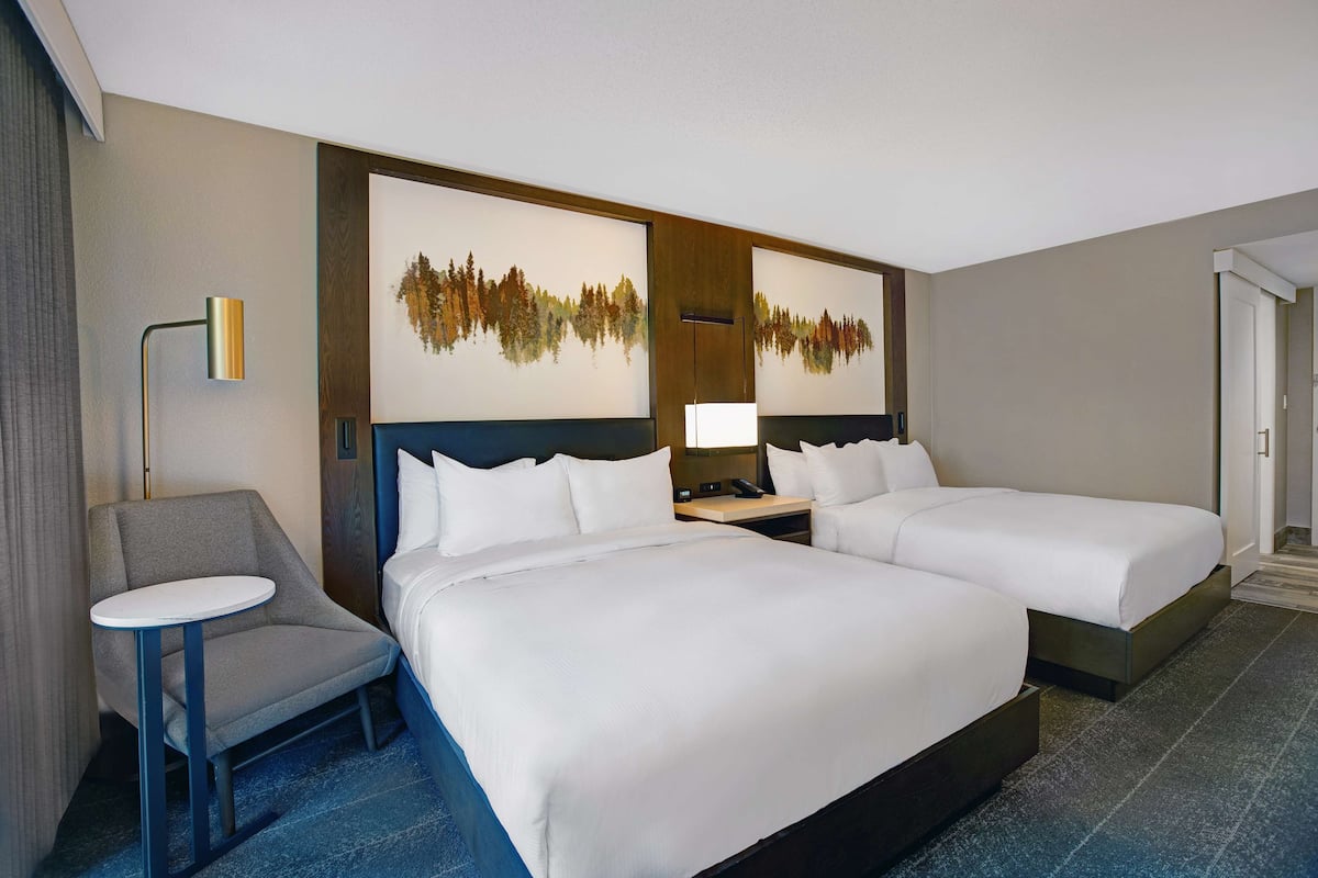 Habitación de hotel con dos camas, una mesita, una silla, una lámpara y cuadros en la pared.