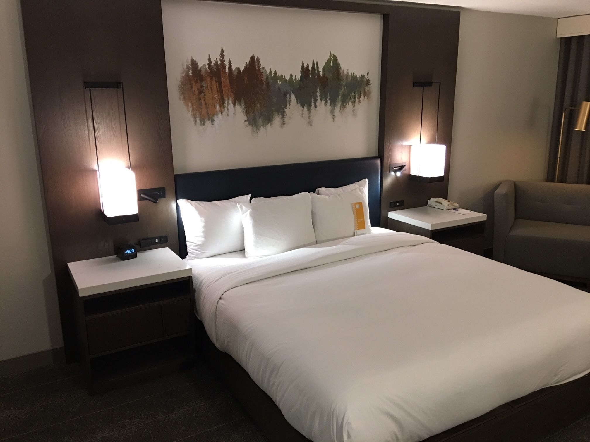 A hotel room with a large bed, two bedside tables with lamps, a nightstand, a chair, and a wall-mounted artwork.