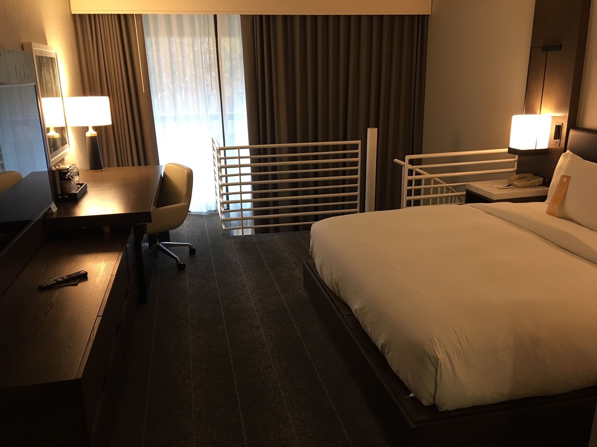 A hotel room with a bed, desk, chair, and a balcony with curtains.