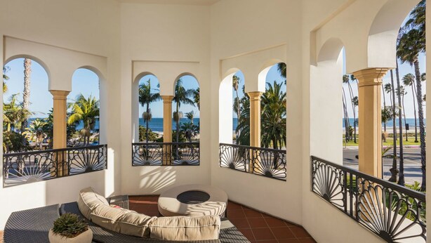 A sunlit balcony with a curved sofa, a small round table, and a potted plant. The balcony features arched windows with a view of palm trees and the sea. - Santa Barbara Inn (Santa Barbara)