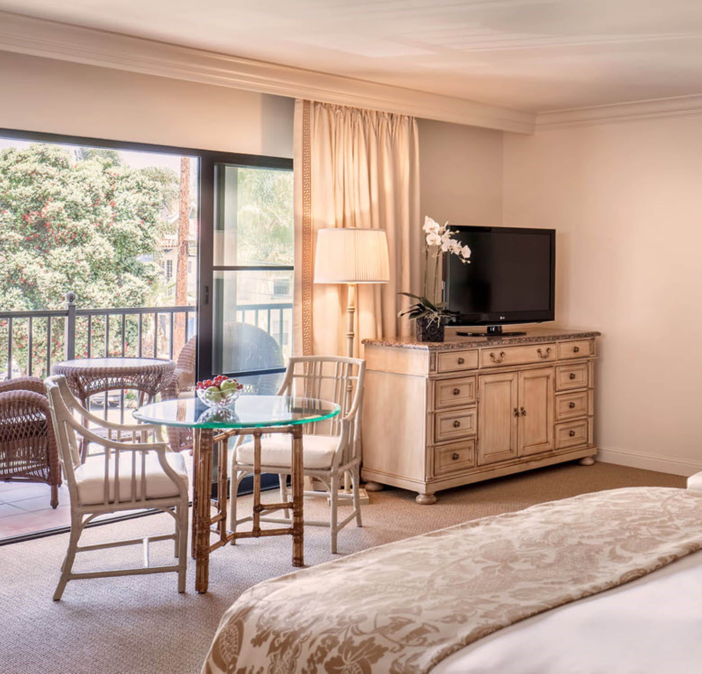 Ein Hotelzimmer mit einem Bett, einem Glastisch mit Stühlen, einem Fernseher auf einer Kommode und einem Balkon mit Blick auf Bäume.