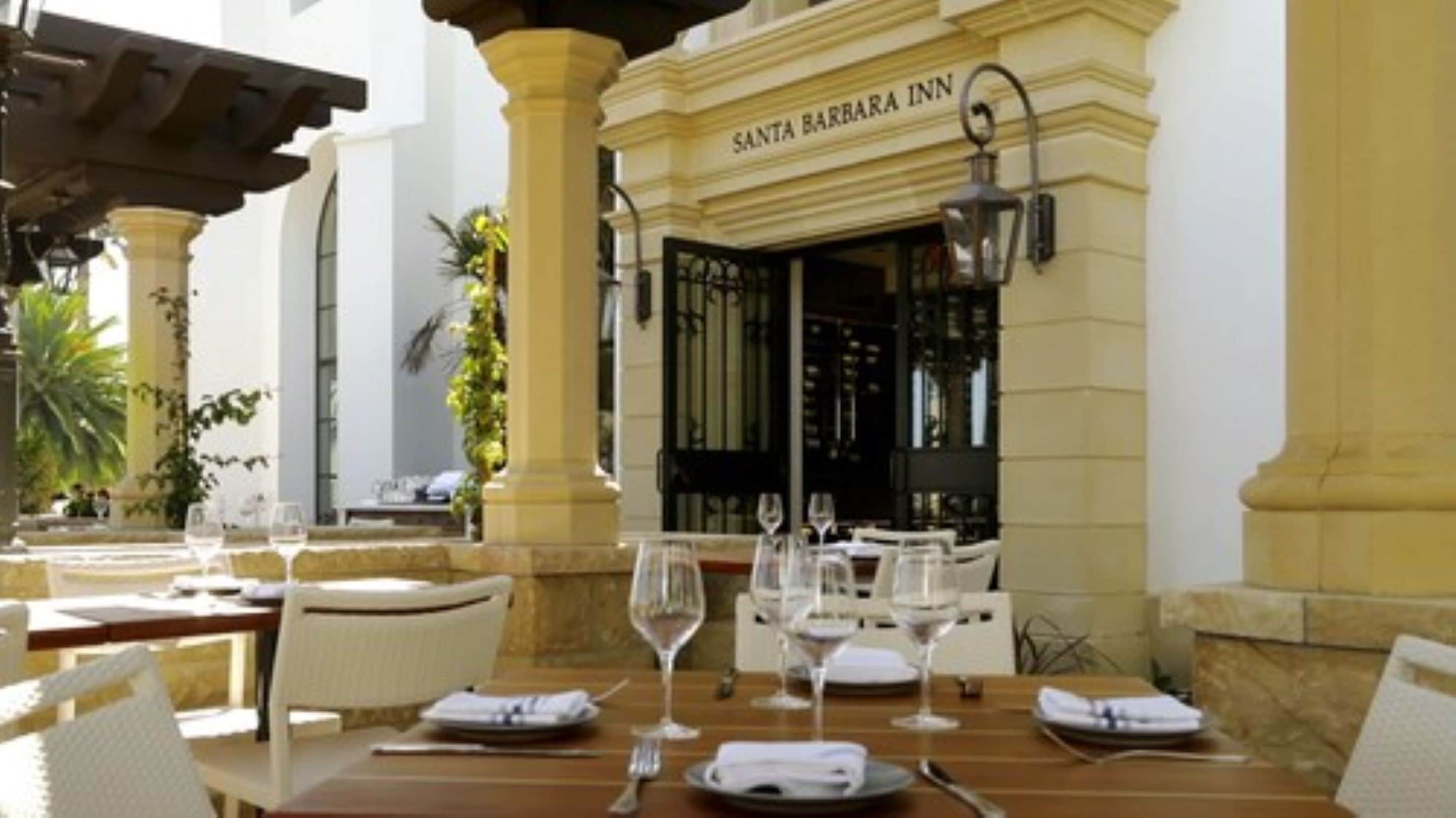 An outdoor dining area at Santa Barbara Inn with tables set for two, featuring white chairs and wine glasses.