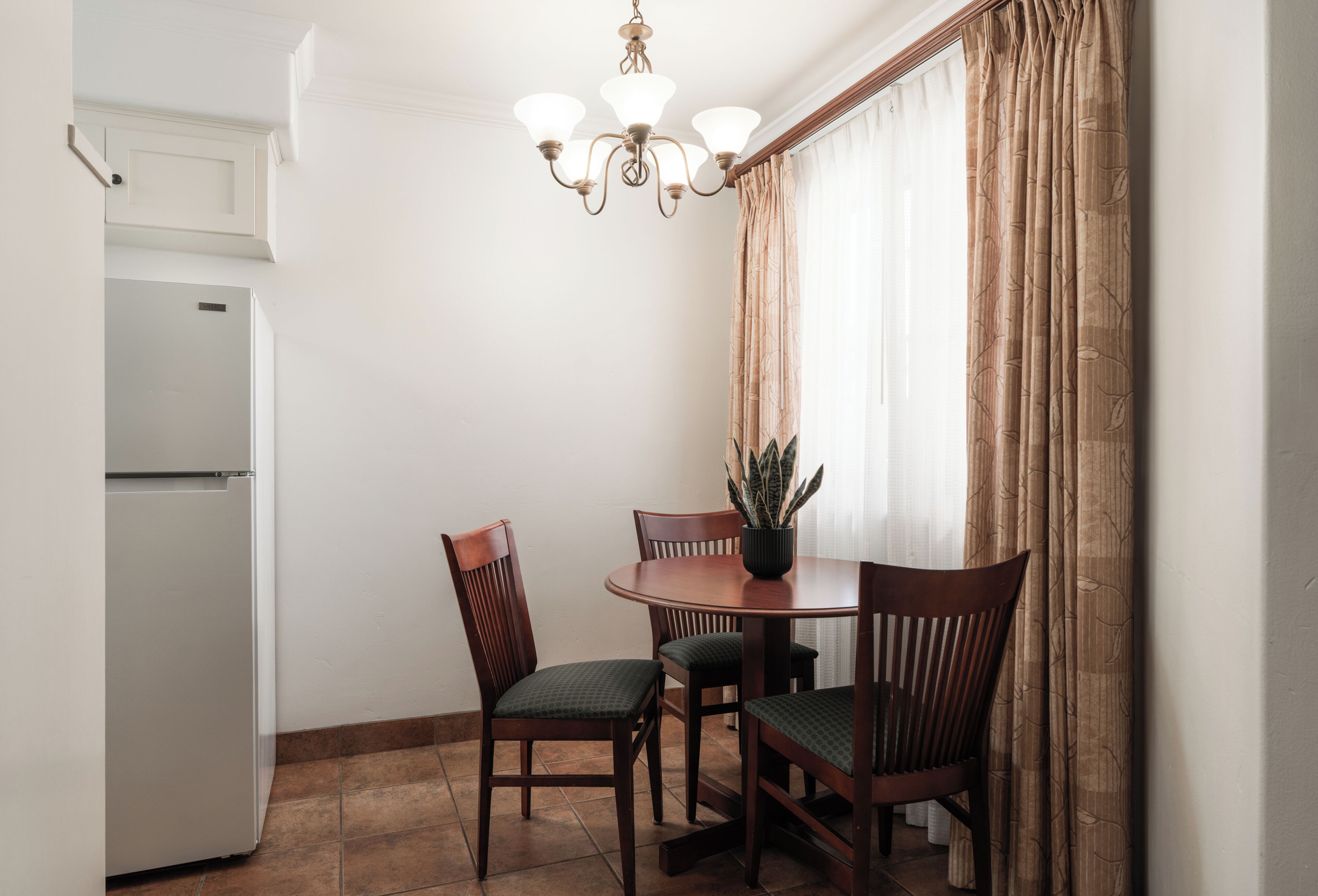 A dining area with a table, chairs, a potted plant, a chandelier, and a refrigerator.