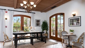A dining area with a wooden table, chairs, and a bench. There are two doors leading to a balcony, and a chandelier hangs from the ceiling. - Brisas del Mar, Inn at the Beach (Santa Barbara)