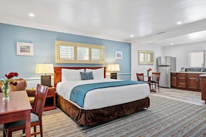 A hotel room with a bed, bedside tables, a desk, chairs, a refrigerator, and a window with blinds.