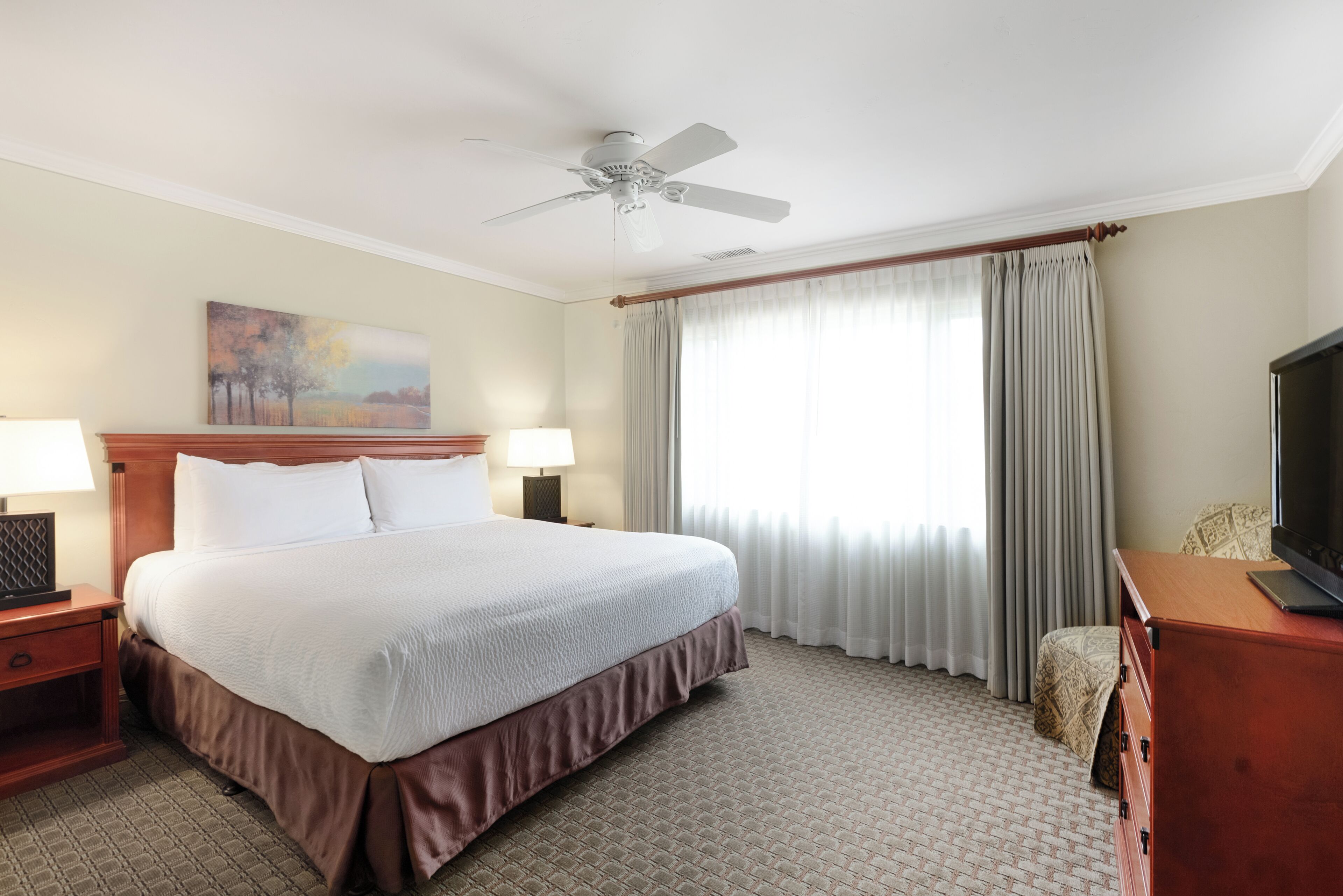 A hotel room with a bed, a ceiling fan, a television, a painting on the wall, and a window with curtains.