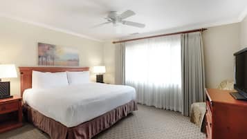 A hotel room with a bed, a ceiling fan, a television, a painting on the wall, and a window with curtains.