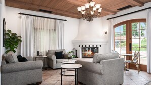 A cozy living room with a fireplace, two armchairs, a round coffee table, and a dining area with a view of the outdoors. - Brisas del Mar, Inn at the Beach (Santa Barbara)