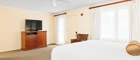 Habitación de hotel con cama, televisor sobre un mueble de madera, ventilador de techo y ventana con cortinas.