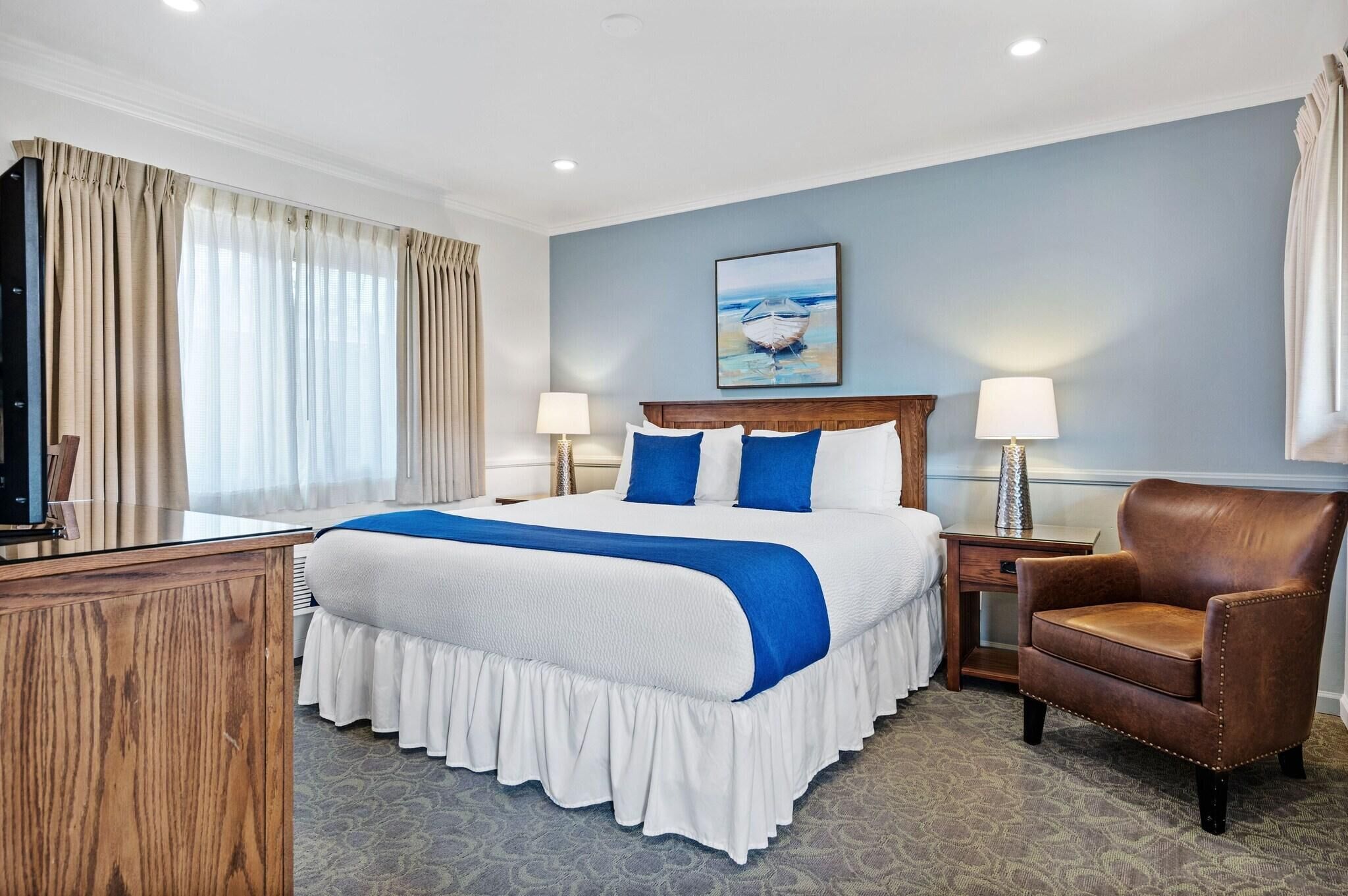 A hotel room with a large bed, two bedside tables with lamps, a wooden desk, a brown leather armchair, and a painting on the wall.