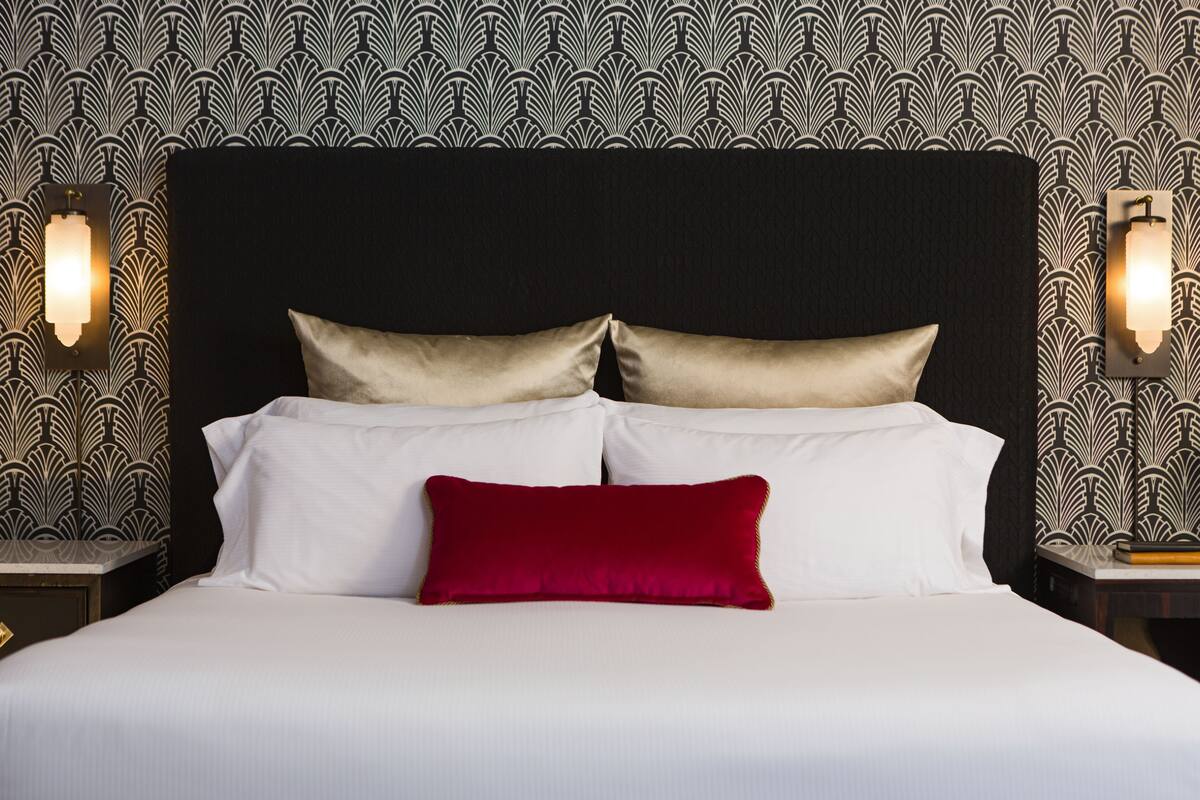 Een net opgemaakt bed met witte lakens en twee gouden kussens, tegen een zwart hoofdeinde met behangpapier met een patroon.