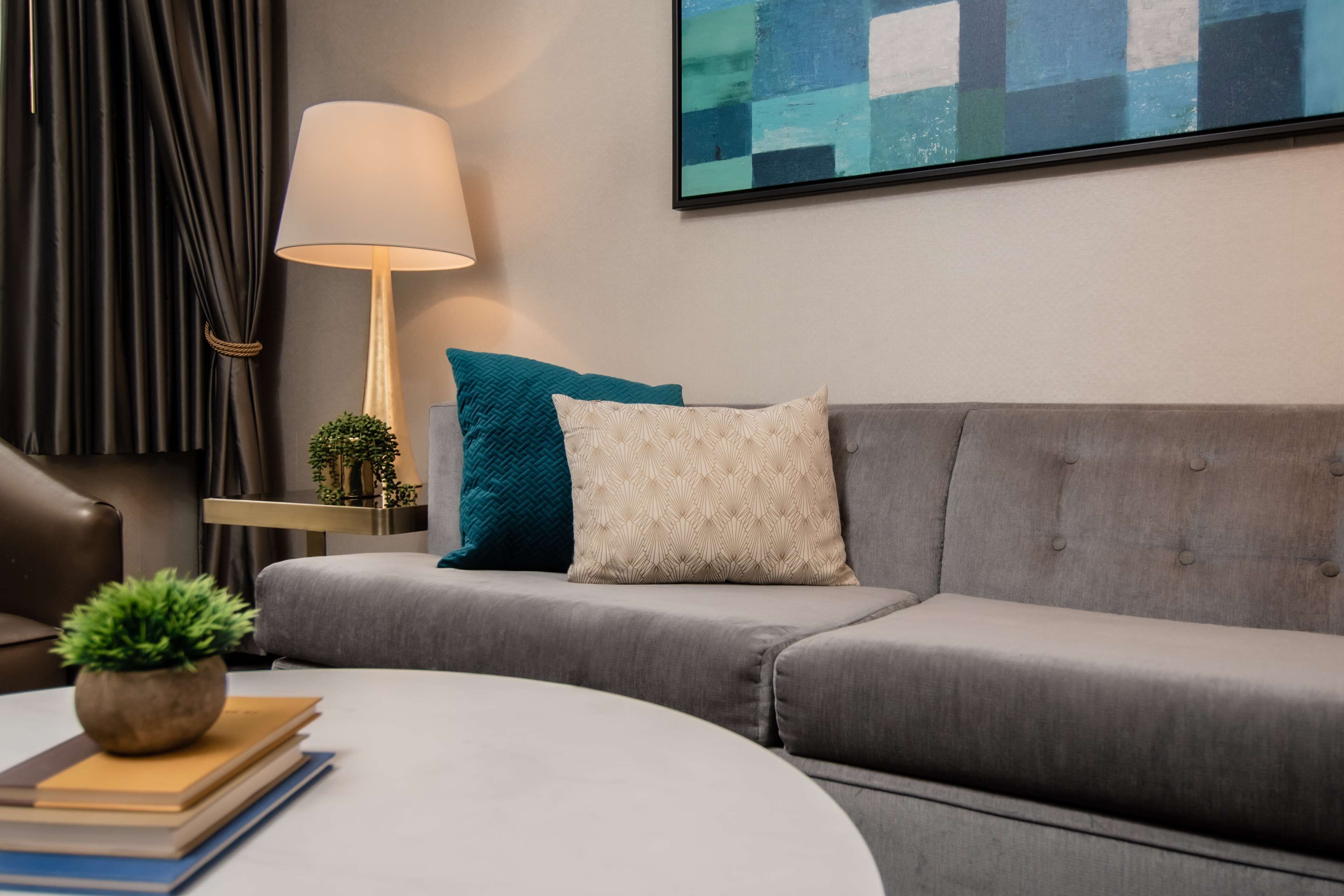 Una sala moderna con un sofá gris, una mesa de centro redonda y una lámpara. Hay una planta sobre la mesa de centro y una mesita de noche con una lámpara y una planta al lado del sofá.