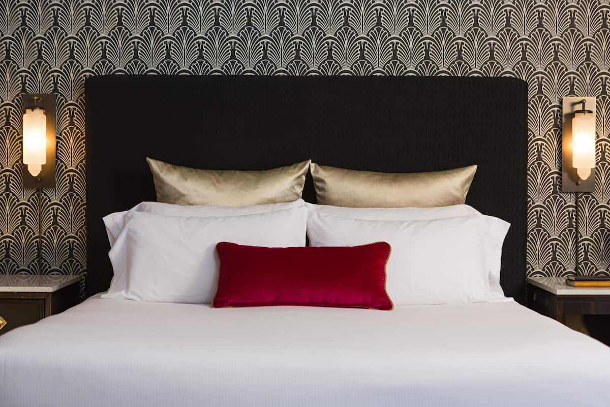 Een bed met witte lakens en twee kussens, één rood en één goud, tegen een behang met patroon.