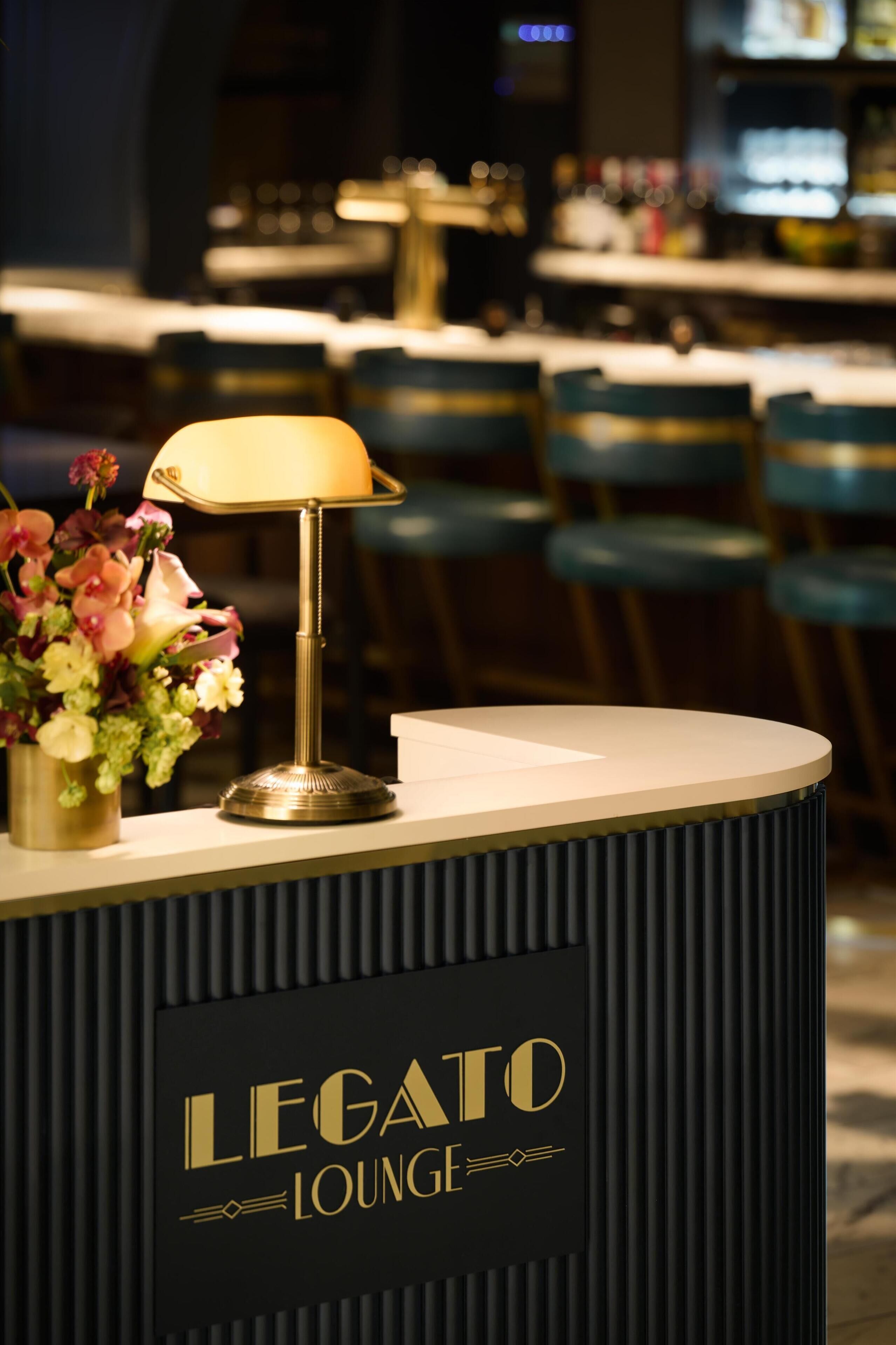 Un comptoir d’accueil agrémenté d’une lampe et de fleurs, portant l’inscription « Legato Lounge ».