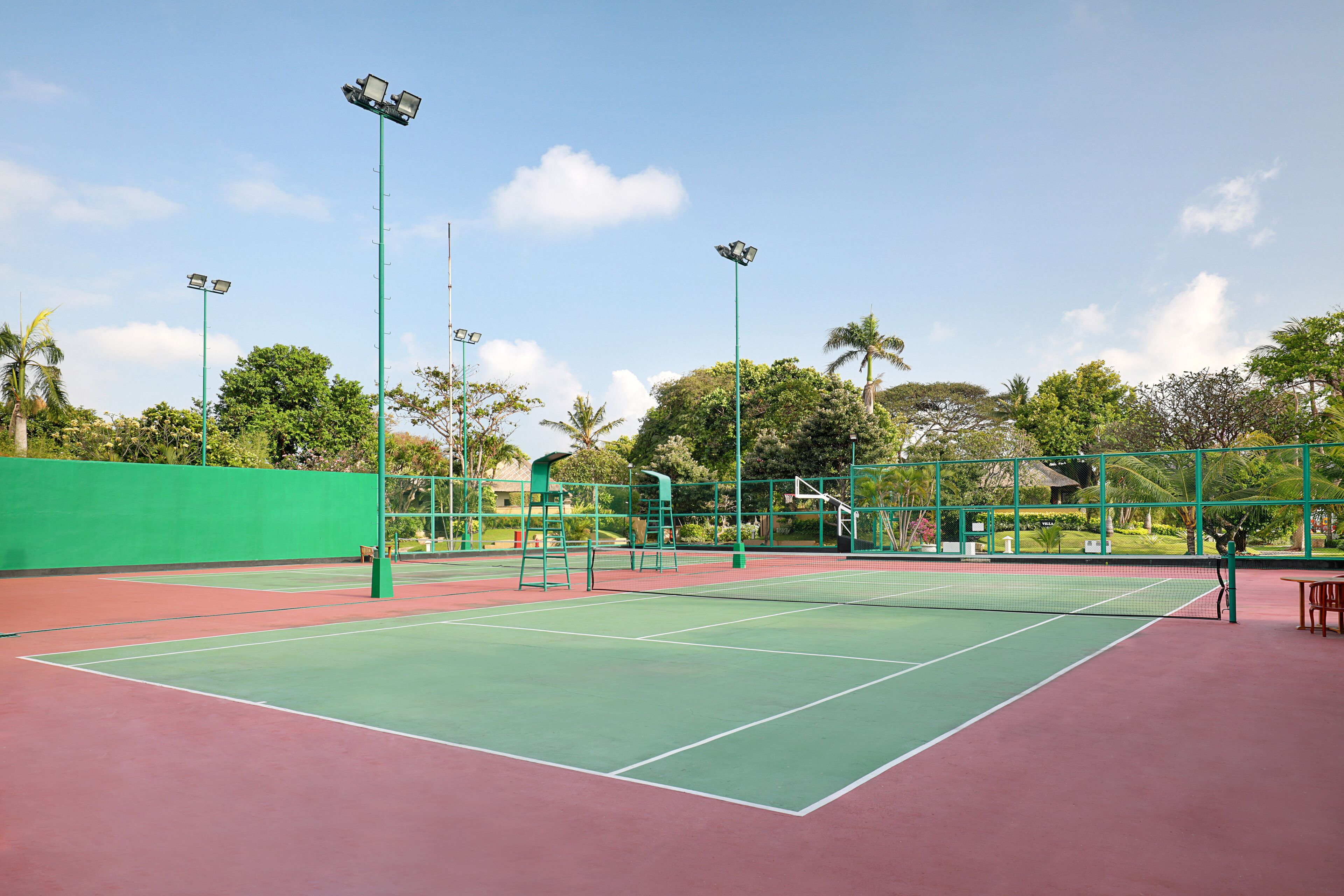 Un court de tennis extérieur avec une surface verte et rouge, entouré d’arbres et d’une clôture verte.