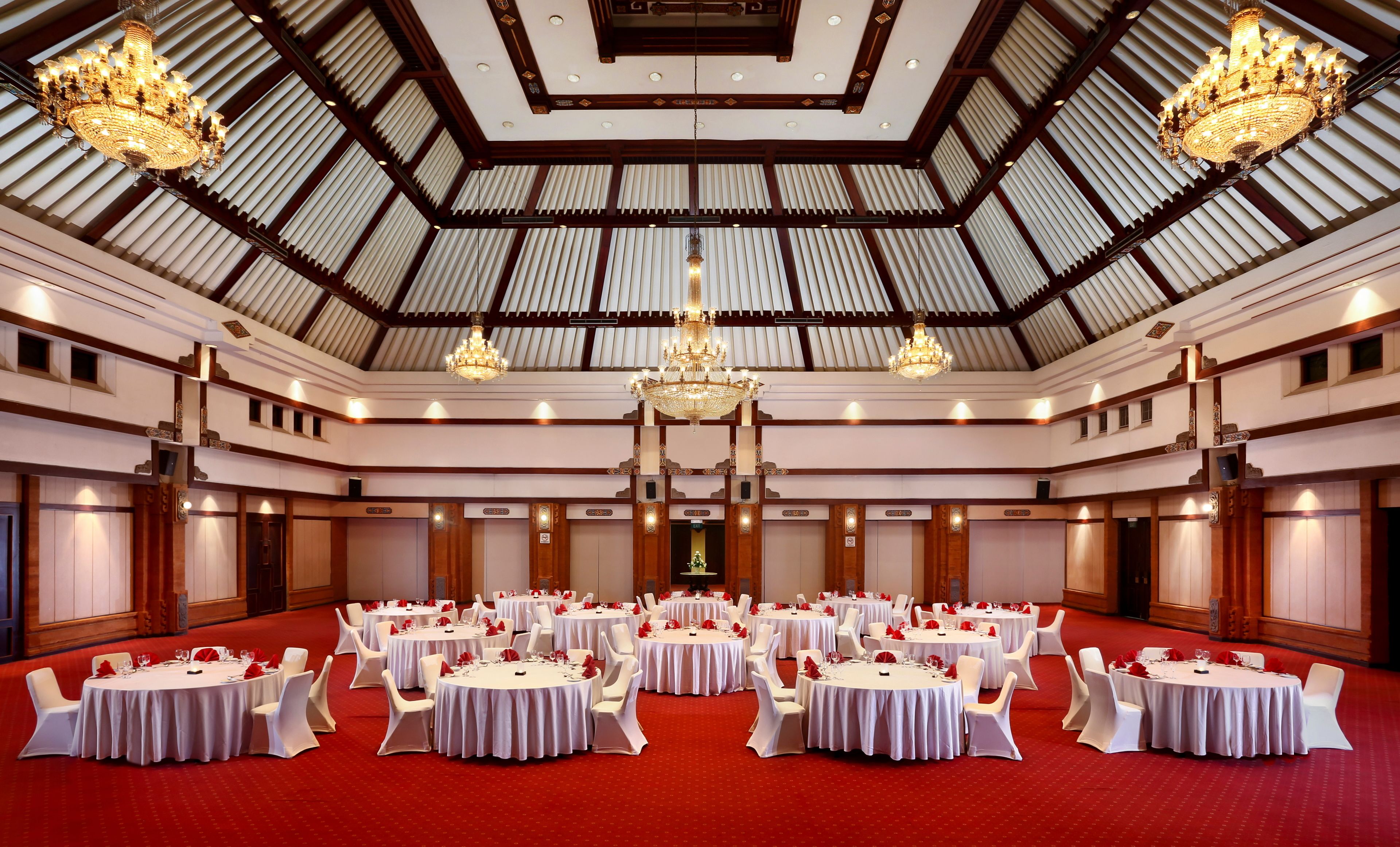 Een ruime balzaal met meerdere ronde tafels, witte tafellakens en rode bloemen als decoratie.
