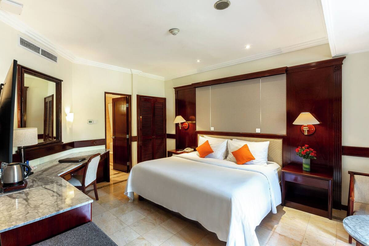 Ein Hotelzimmer mit einem großen Bett, einem Schreibtisch mit Stuhl, einem Spiegel, einem Nachttisch mit Lampe und einem kleinen Tisch mit Blumen.