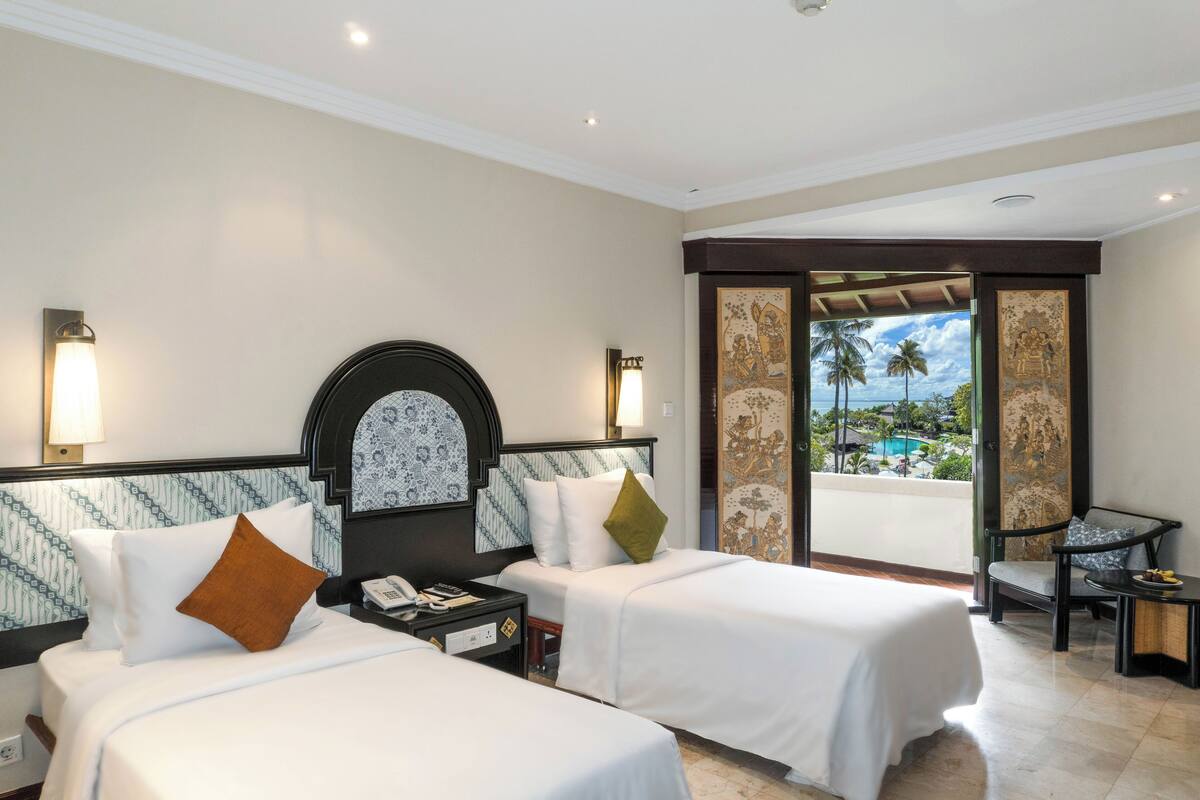 Ein Hotelzimmer mit zwei Betten, einem Nachttisch, einem Sessel und Blick auf einen Pool und Palmen.