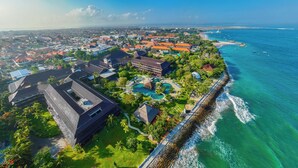 Vista aérea de un complejo turístico costero con una extensa playa, una piscina y un rompeolas rocoso.
