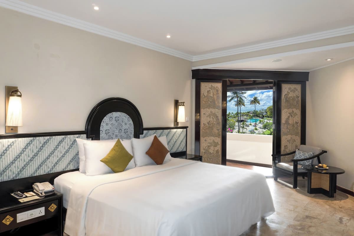 Ein Hotelzimmer mit einem großen Bett, einem Nachttisch mit Telefon, einem Sessel und einem kleinen Tisch mit Obstteller.