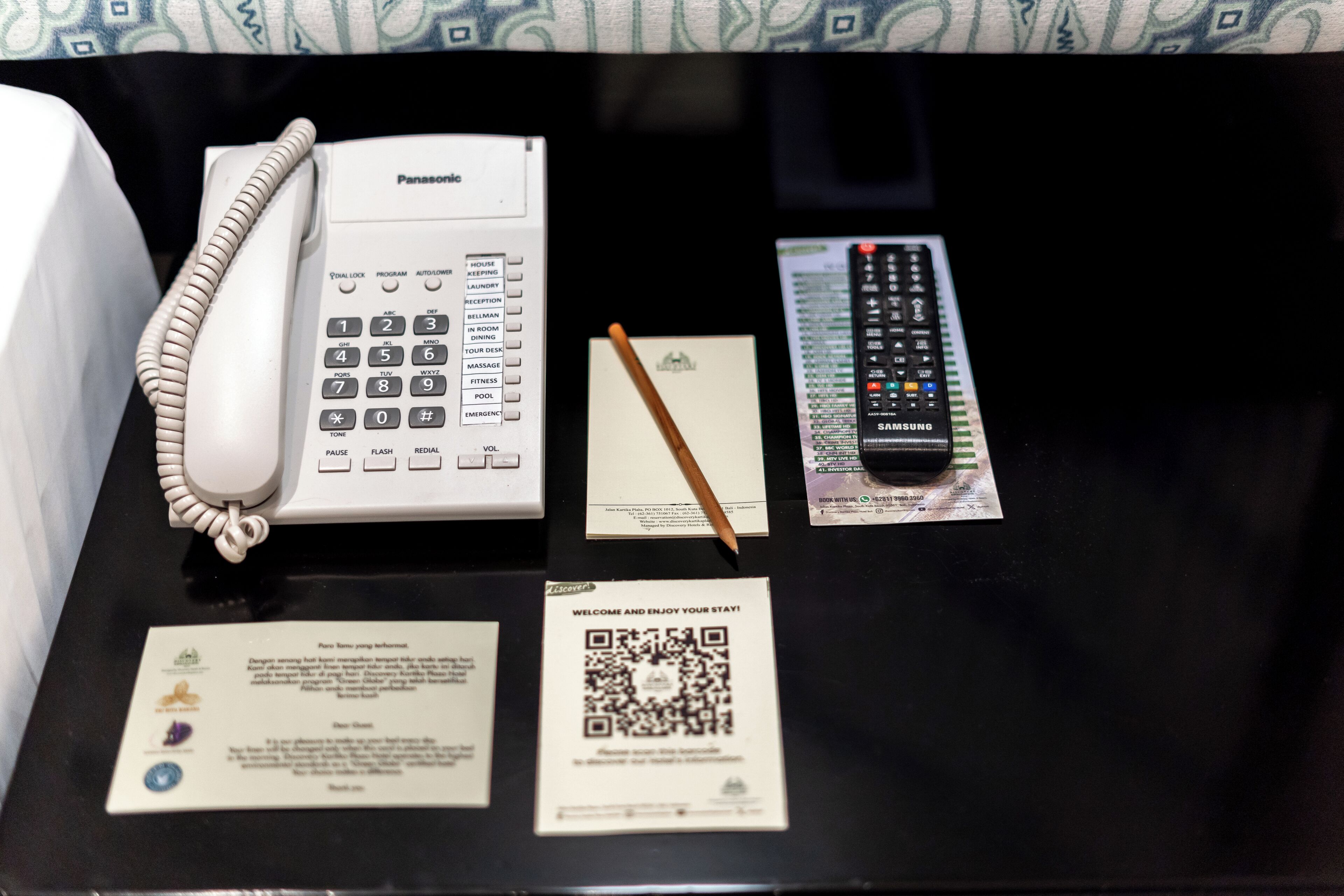 A hotel room with a Panasonic telephone, a Samsung remote control, a notepad, a pen, and a welcome card.