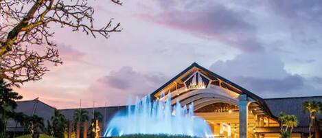 Un hotel con una fuente en primer plano, un paseo cubierto y un cielo al atardecer.