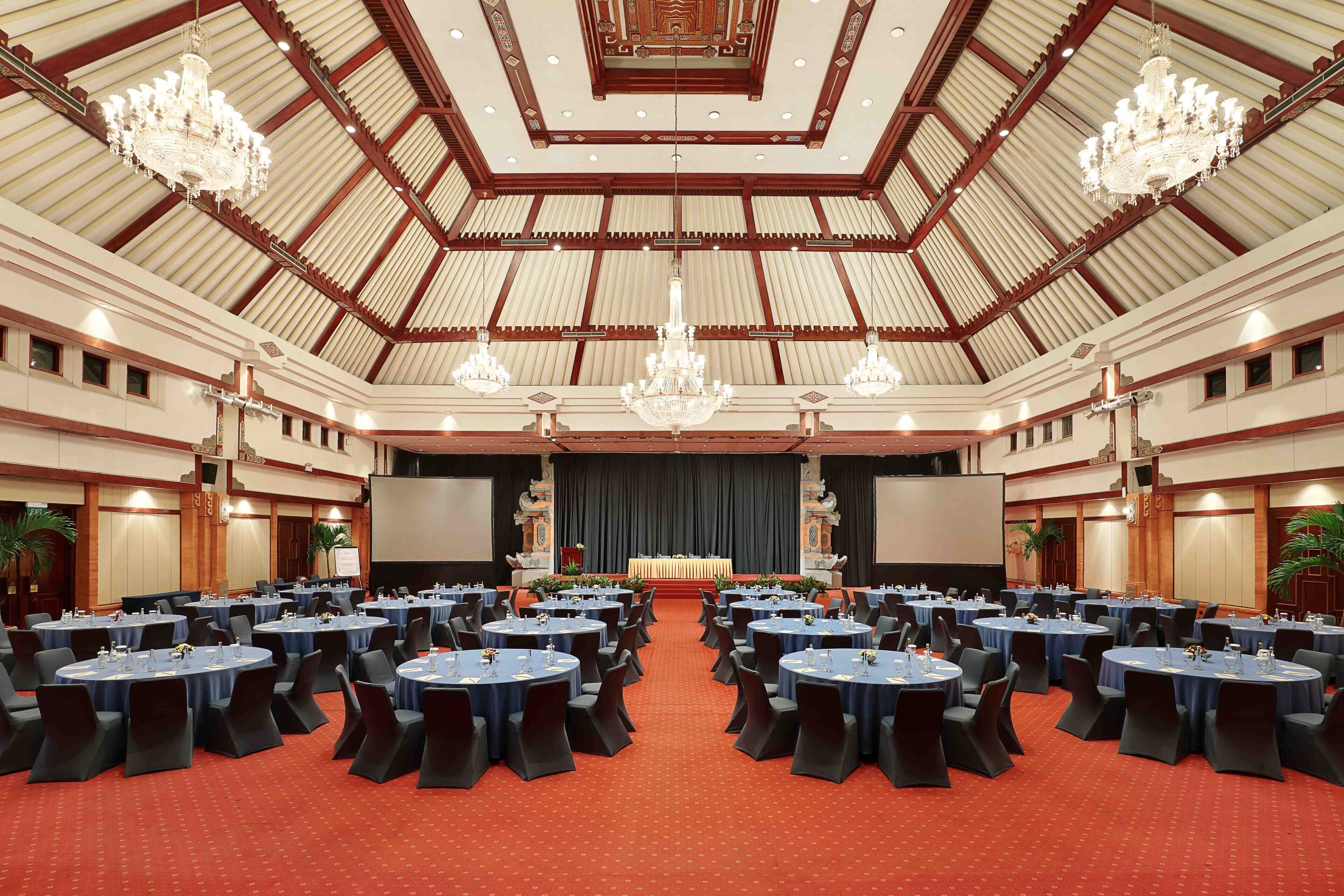Un salón de conferencias amplio con mesas y sillas redondas, candelabros y un escenario con una pantalla.