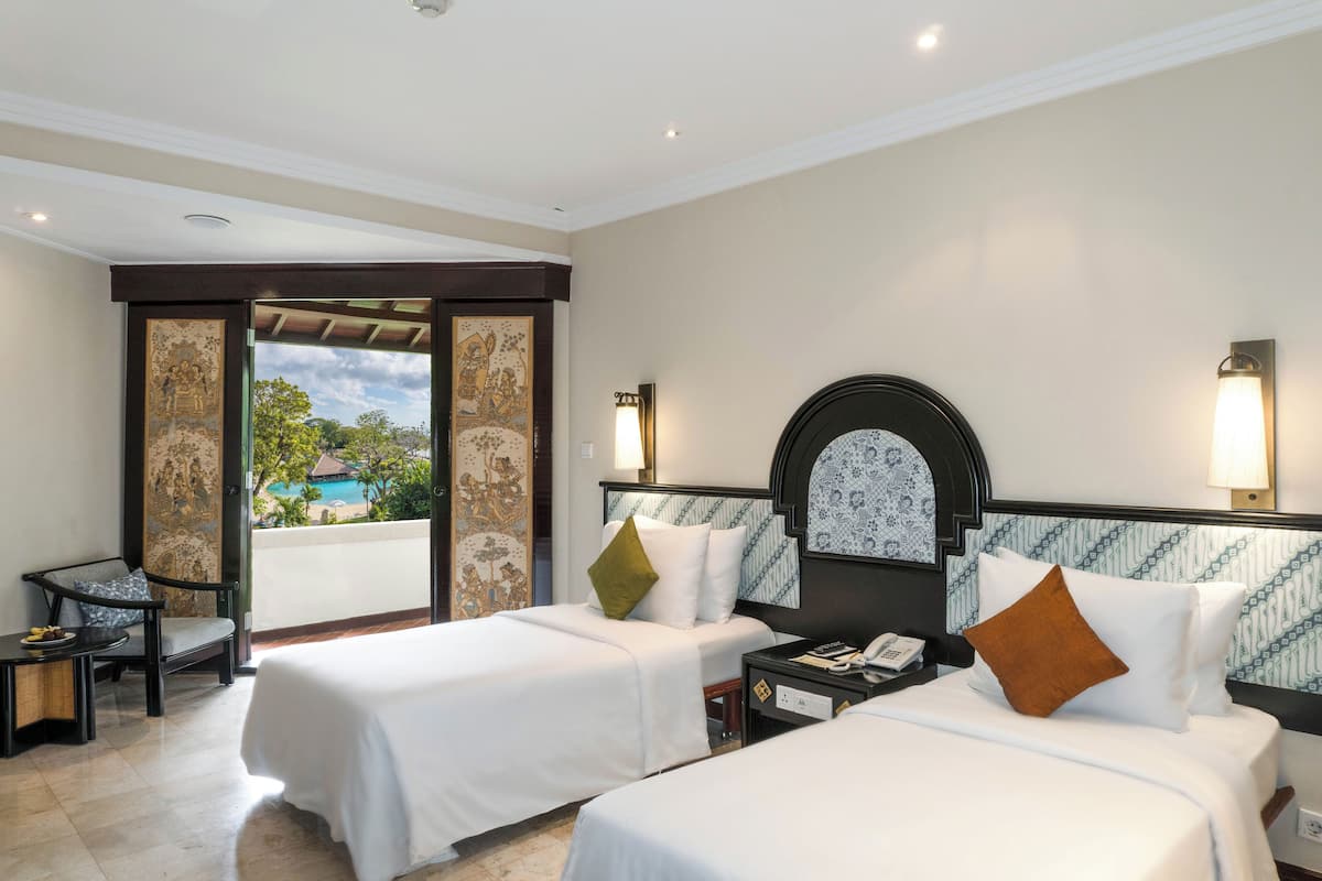 Ein Hotelzimmer mit zwei Betten, einem kleinen Tisch, einem Sessel und Blick auf einen Pool und Bäume.