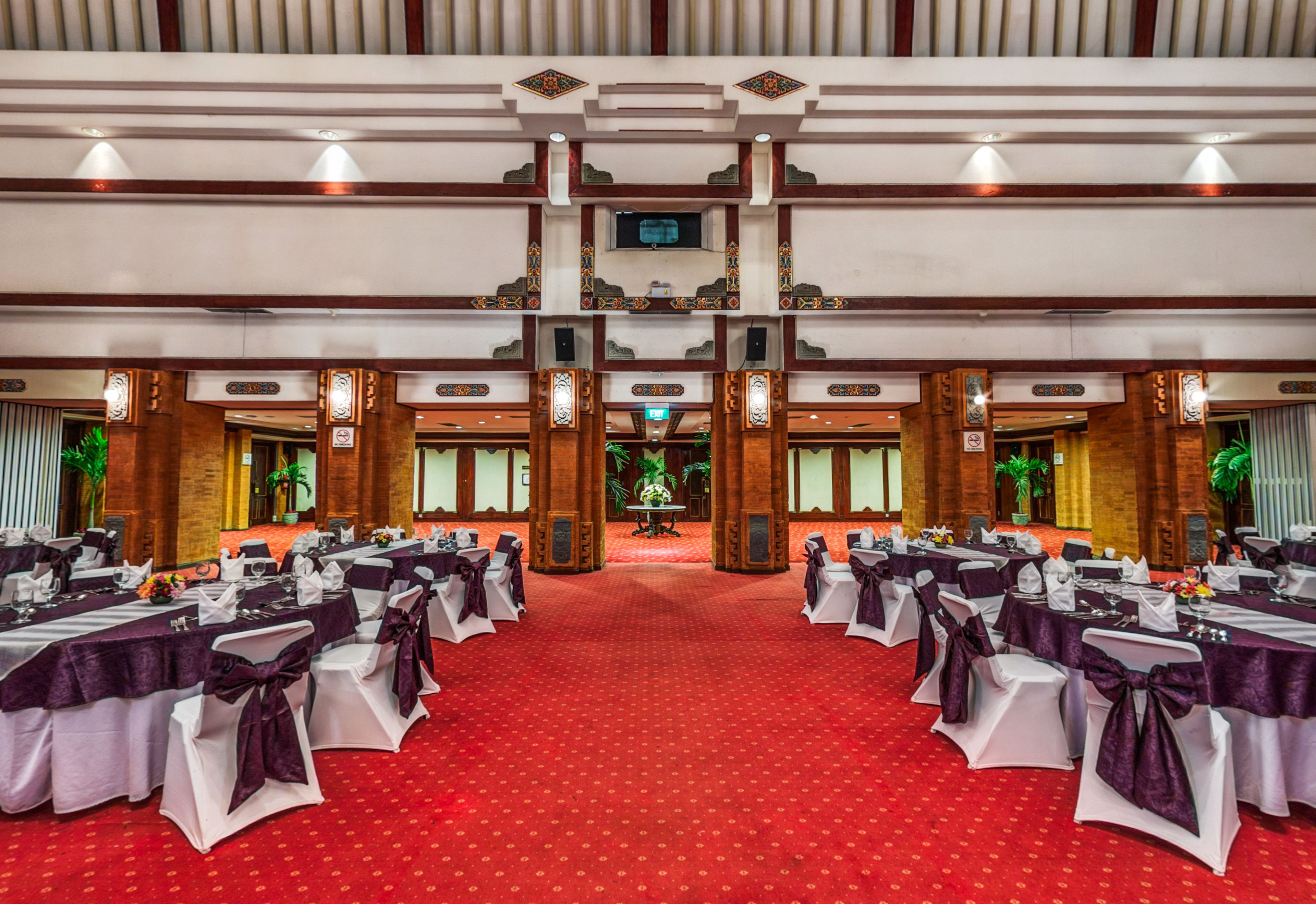 Une salle de banquet avec des tables disposées en rangées, un tapis rouge et un plafond haut orné d’éléments décoratifs.