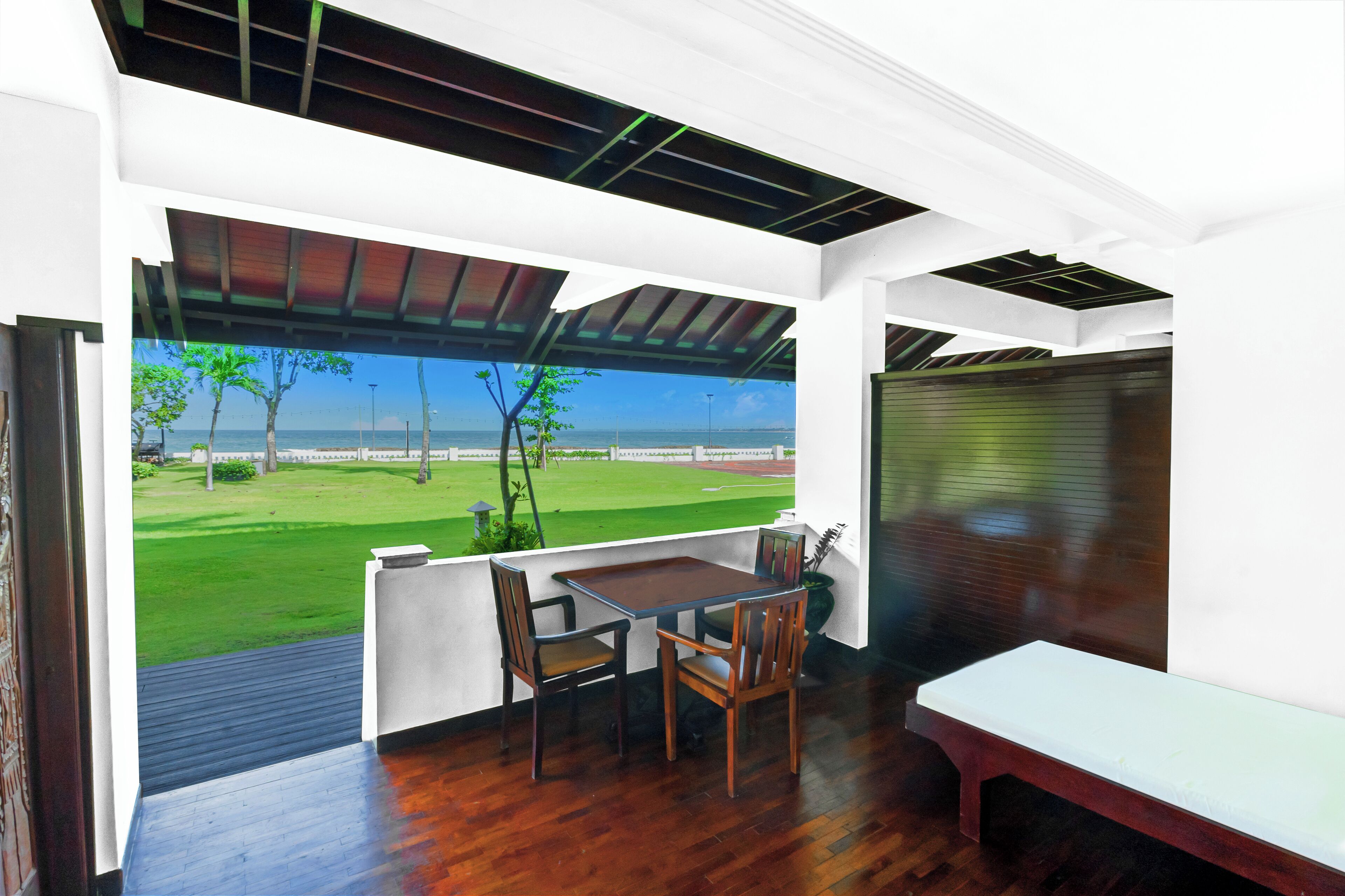 Quarto moderno com piso de madeira, um banco branco e uma mesa com cadeiras. Uma janela grande oferece vista para um gramado verde e árvores.