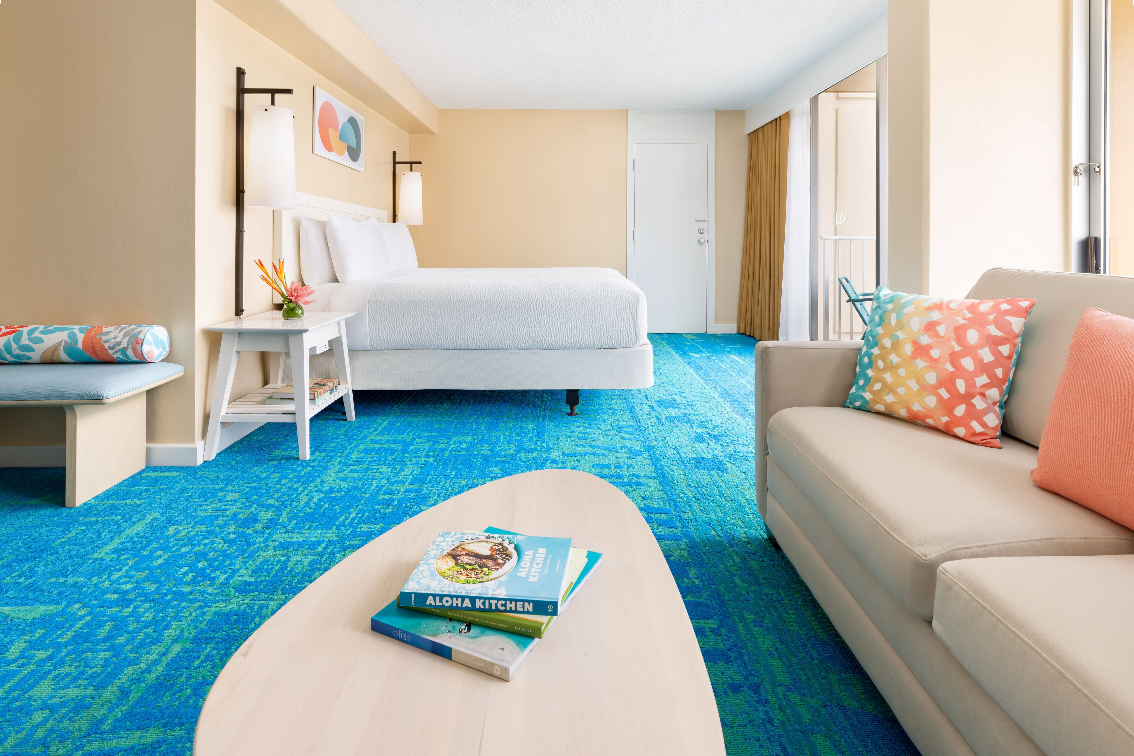 Habitación de hotel con cama, sofá, mesa de centro con un libro, mesita de noche y ventana con cortinas.