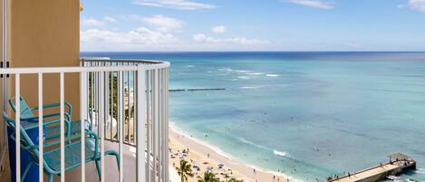A balcony with turquoise chairs, a view of a beach with clear turquoise water, and a pier extending into the ocean.