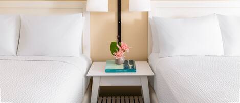 A hotel room with two beds, a small bedside table with a book and a vase of flowers, and two wall-mounted lamps.