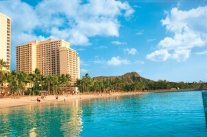 Un hotel frente a la playa con piscina, palmeras y una montaña al fondo.
