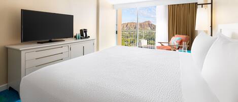 A hotel room with a large bed, a flat-screen TV, a desk with a coffee maker, and a balcony with a view of mountains.